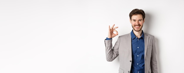 Free photo smiling successful businessman in suit showing okay sign looking satisfied standing against white ba