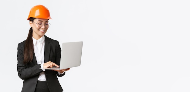 Улыбающаяся успешная азиатская женщина-инженер-менеджер фабрики в защитном шлеме и деловом костюме с помощью портативного компьютера проверяет проект или чертежи на экране