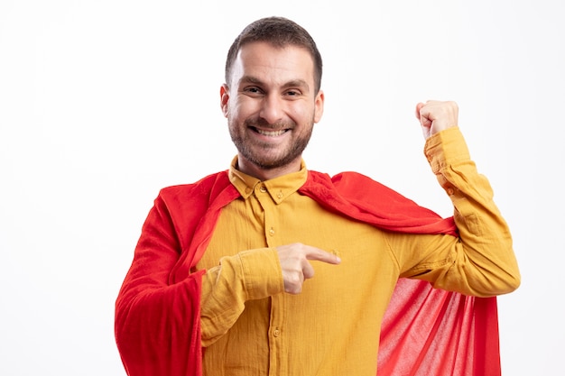 Smiling superhero man with red cloak tenses and points at biceps isolated on white wall