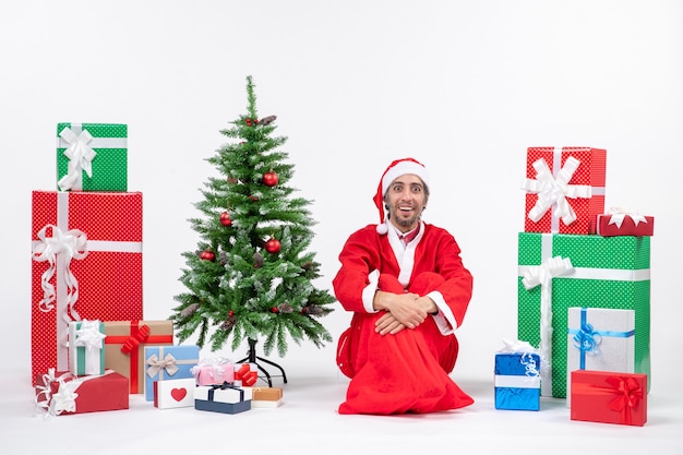 Sorridente sorpreso giovane uomo vestito da babbo natale con doni e albero di natale decorato seduto per terra su sfondo bianco