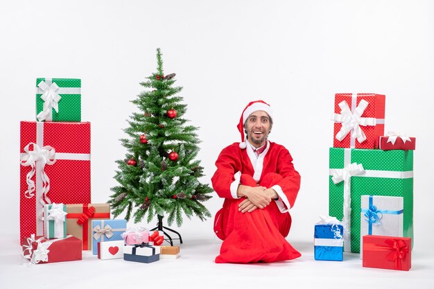 Smiling surprised young man dressed as Santa claus with gifts and decorated Christmas tree sitting on the ground on white background