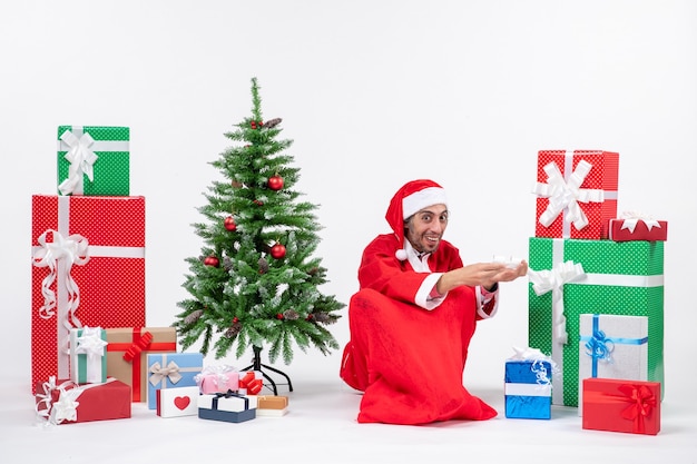 Улыбающийся удивленный молодой человек в костюме Санта-Клауса с подарками и украшенной елкой сидит на земле на белом фоне