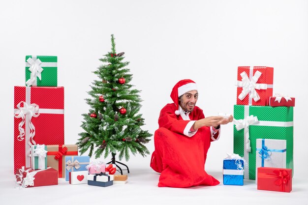 Smiling surprised young man dressed as Santa claus with gifts and decorated Christmas tree sitting on the ground on white background