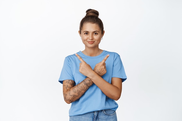 Free photo smiling tattooed girl pointing fingers sideways, showing two options, standing in blue t-shirt on white