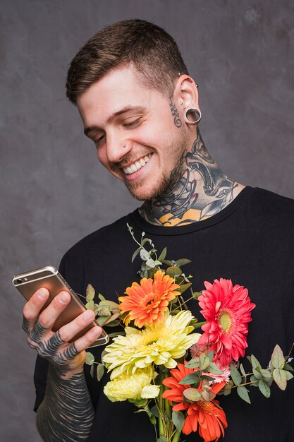 Smiling tattooed young man with pierced ears and nose holding bouquet using smartphone