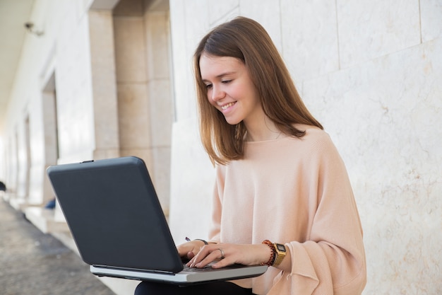 Smiling teen girl browsing on laptop on bench at building