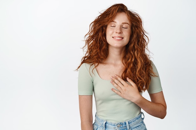 Foto gratuita sorridente ragazza adolescente con i capelli rossi pensando a qualcosa con gli occhi chiusi tenendo la mano sul cuore sognare ad occhi aperti scena commovente scena sfondo bianco