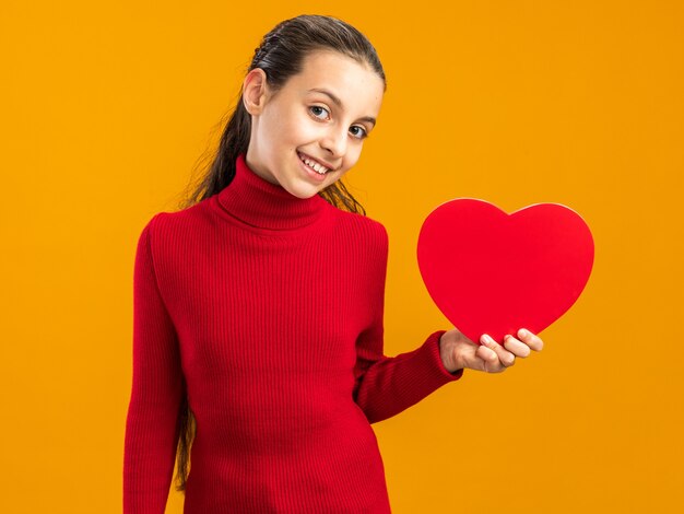 Smiling teenage girl holding heart shape looking at front isolated on orange wall