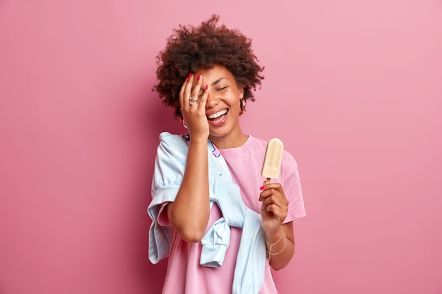 Foto gratuita l'adolescente sorridente rende il palmo del viso felice di indossare una maglietta casual con il maglione si gode l'estate mangia un delizioso gelato sul bastone ha buon umore isolato sul muro rosa. divertimento e dolci