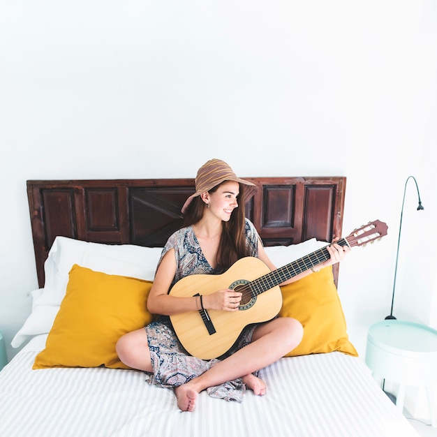 Smiling teenage girl sitting on bed playing guitar in bedroom