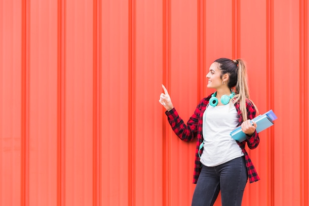 Smiling teenage girl standing against an orange corrugated wall pointing her finger at something