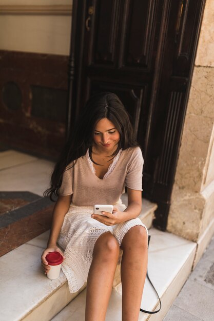 Smiling teenage girl using mobile phone holding takeaway coffee cup