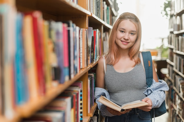 Улыбаясь подросток с книгой, опираясь на книжный шкаф