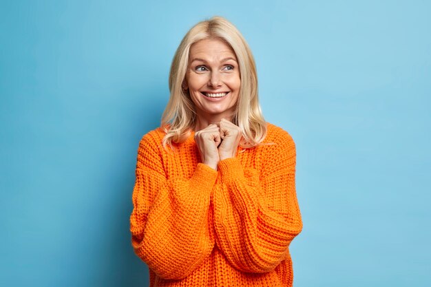 smiling tender middle aged woman with light hair toothy smile keeps hands together and looks away has dreamy expression dressed in oversized jumper stands over blue wall.