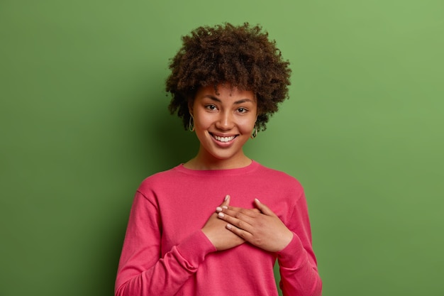 Smiling tender woman presses hands to chest in grateful thankful gesture, appreciates good words and expresses gratitude, feels flattered to receive romantic gift, isolated on green wall.