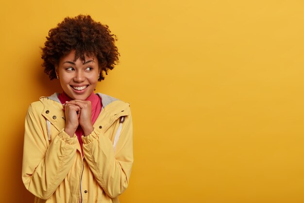 Smiling tender young woman keeps hands under chin, gazes aside with broad toothy smile, wears casual anorak, dreams about something pleasant, poses against yellow  wall, empty space area