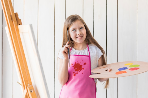 Smiling thoughtful girl holding palette and paint brush standing near the easel