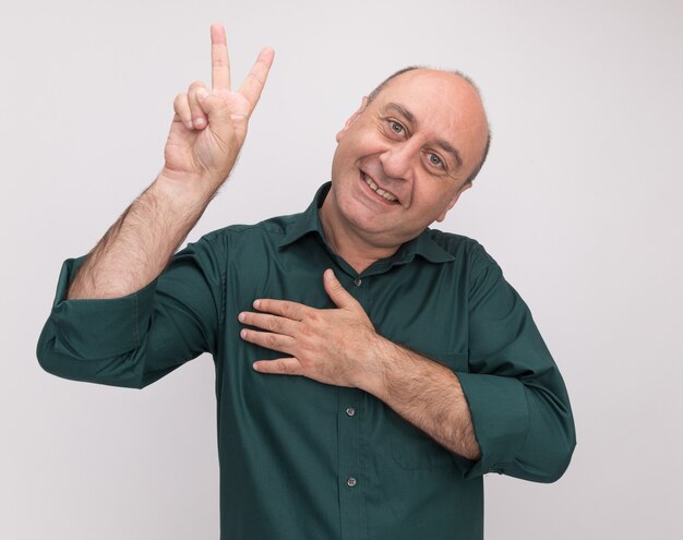 Smiling tilting head middle-aged man wearing green t-shirt showing peace gesture putting hand on heart isolated on white wall