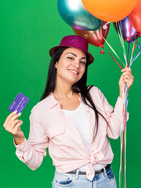 Smiling tilting head young beautiful girl wearing party hat holding balloons with credit card isolated on green wall