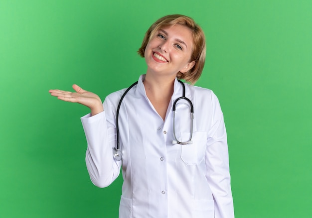 Улыбаясь, наклонив голову, молодая женщина-врач в медицинском халате со стетоскопом указывает рукой сбоку, изолированной на зеленом фоне