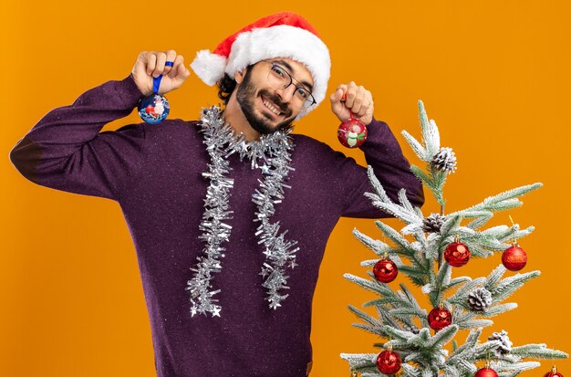 smiling tilting head young handsome guy standing nearby christmas tree wearing christmas hat with garland on neck holding christmas balls isolated on orange wall