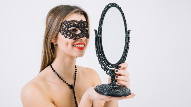 手の鏡で見ているカーニバルマスクでトップレスの女性の笑みを浮かべてください。