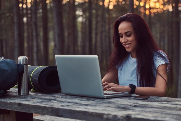 Улыбающаяся туристка-хипстерша в белой рубашке работает на ноутбуке, сидя на деревянной скамейке в красивом осеннем лесу.