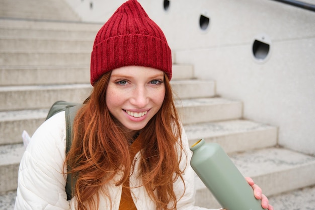 Бесплатное фото Улыбающаяся путешественница рыжеволосая туристка сидит на лестнице с флягой и пьет горячий кофе из термоса, пока