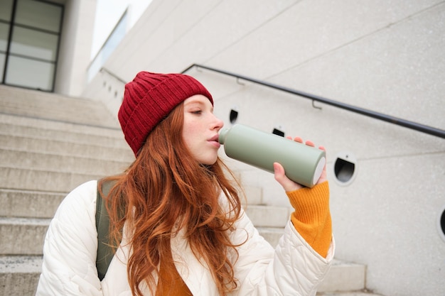 Free photo smiling traveler redhead girl tourist sits on stairs with flask drinks hot coffee from thermos while