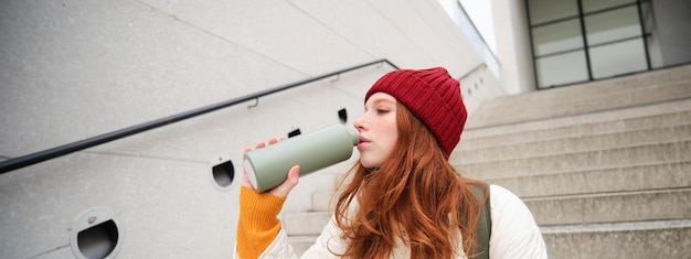 Free photo smiling traveler redhead girl tourist sits on stairs with flask drinks hot coffee from thermos while