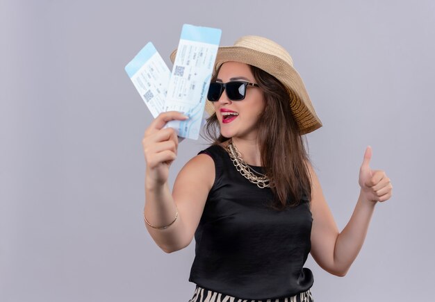 Smiling traveler young girl wearing black undershirt in hat in gkasses holdings tickets her thumb up on white background