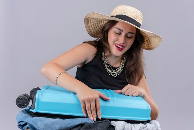 Улыбающаяся молодая девушка путешественника в черной майке в шляпе держит открытый чемодан на белом фоне