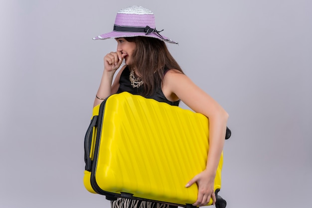 Smiling traveler young girl wearing black undershirt in hat holding suitcase on white background