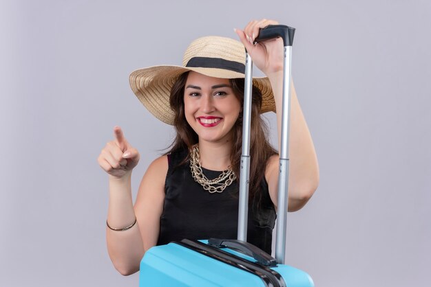Улыбающаяся молодая девушка путешественника в черной майке в шляпе положила руку на чемодан и указывает в сторону на белом фоне