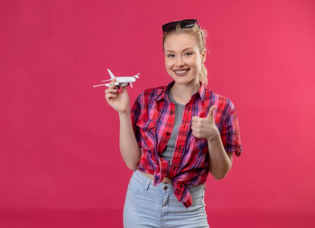 Smiling traveler young girl wearing red shirt and glasses on her head holding toy plan her thumb up on isolated pink background