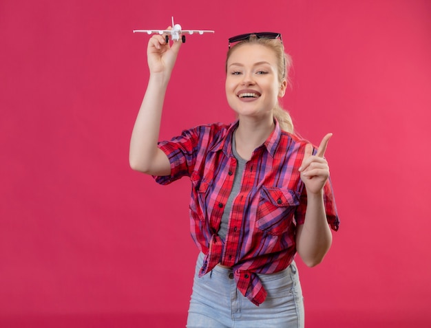 Smiling traveler young girl wearing red shirt and glasses on her head playing with toy plan points to up on isolated pink background