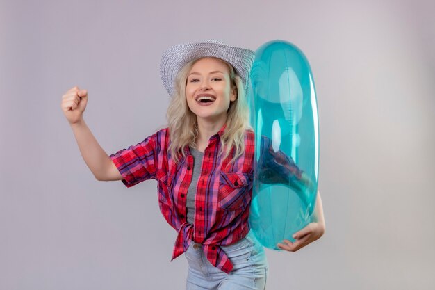 Smiling traveler young girl wearing red shirt in hat holding inflatable ring doing strong gesture on isolated white background