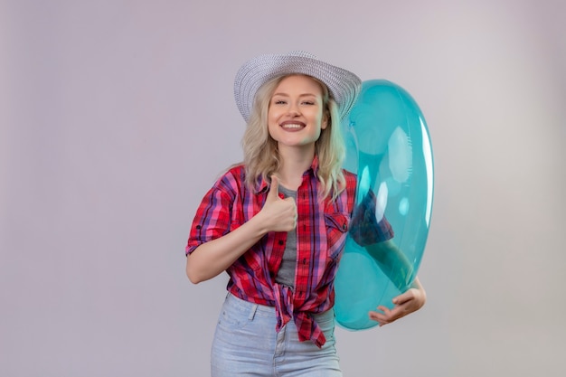 Smiling traveler young girl wearing red shirt in hat holding inflatable ring her thumb up on isolated white background