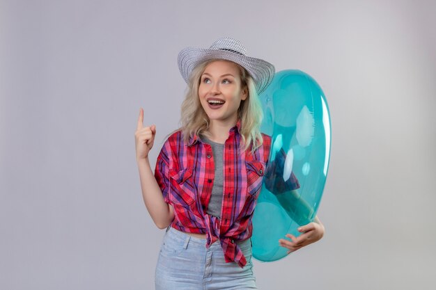 Foto gratuita la ragazza sorridente del viaggiatore che porta la camicia rossa in cappello che tiene i punti gonfiabili dell'anello verso l'alto su fondo bianco isolato