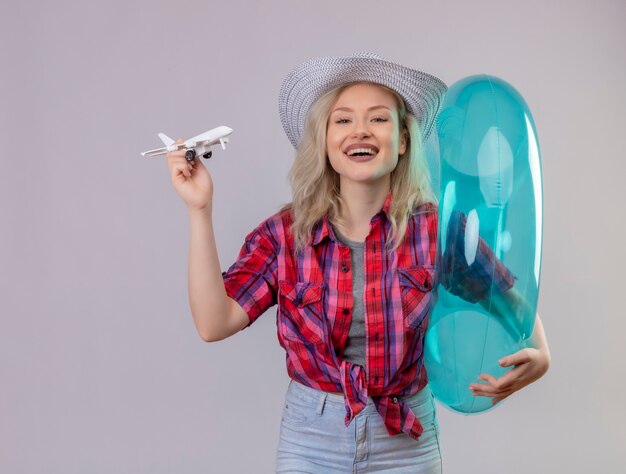 Sorridente ragazza giovane viaggiatore indossa una camicia rossa in cappello che tiene anello gonfiabile e piano giocattolo su sfondo bianco isolato