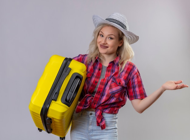 Улыбающаяся молодая девушка путешественника в красной рубашке в шляпе, держащая чемодан, указывает в сторону на изолированном белом фоне