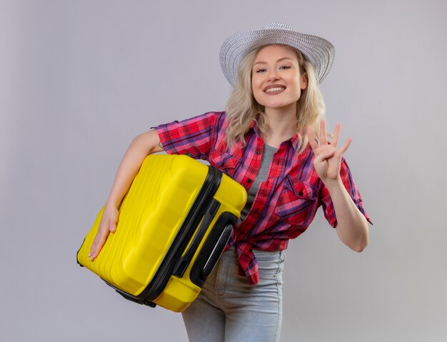 Улыбающаяся молодая девушка путешественника в красной рубашке в шляпе держит чемодан, показывая четыре на изолированном белом фоне