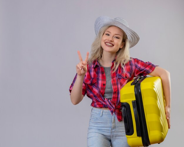 Улыбающаяся молодая девушка путешественника в красной рубашке в шляпе держит чемодан, показывая жест мира на изолированном белом фоне