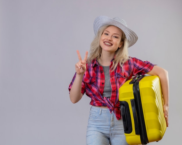 孤立した白い背景の上の平和のジェスチャーを示すスーツケースを持って帽子に赤いシャツを着て旅行者の少女の笑顔
