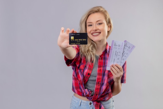 Smiling traveler young girl wearing red shirt holding credit card and tickets on isolated white background