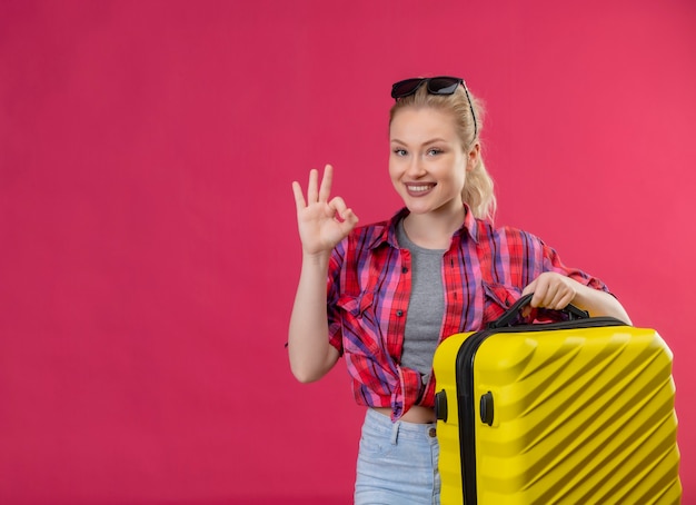 スーツケースを持って赤いシャツを着て笑顔の旅行者の若い女の子は孤立したピンクの背景にオーカージェスチャーを示しています