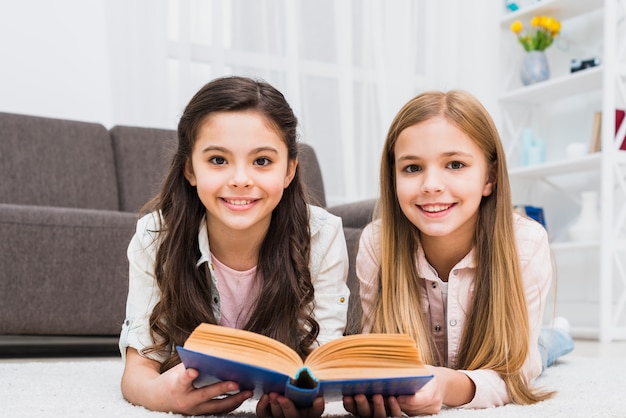 Smiling two female friends lying on carpet holding book in hand