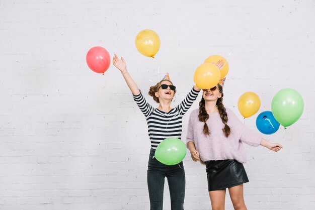 Smiling two female friends standing against white wall playing with colorful balloons