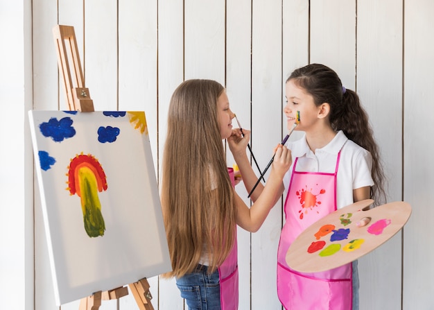 Smiling two girls painting each other face with paint brush standing near the canvas