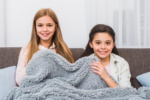 Smiling two girls sitting on sofa covering with grey blanket